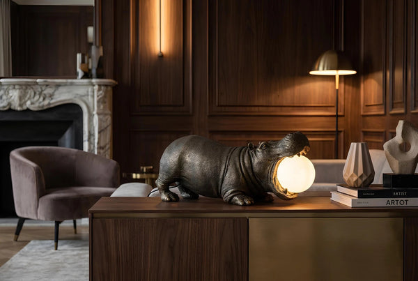 Hippopotamus-shaped lamp on a wooden table in a stylish room with a fireplace and armchair.